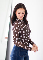 A woman wearing a brown long-sleeve mock neck top with white polka dots, paired with blue jeans, standing against a bright white background.