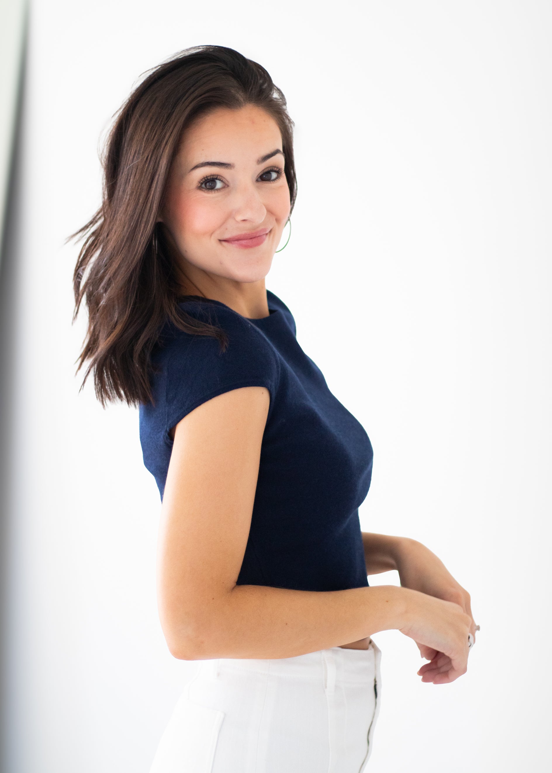 A woman with shoulder-length brown hair is shown from the waist up, wearing a dark navy blue short-sleeve top and white pants against a bright white background.