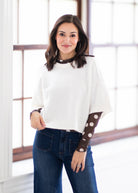 A woman modeling a white knit sweater with long brown sleeves featuring white polka dots, paired with dark blue jeans, standing in a bright room with large windows.