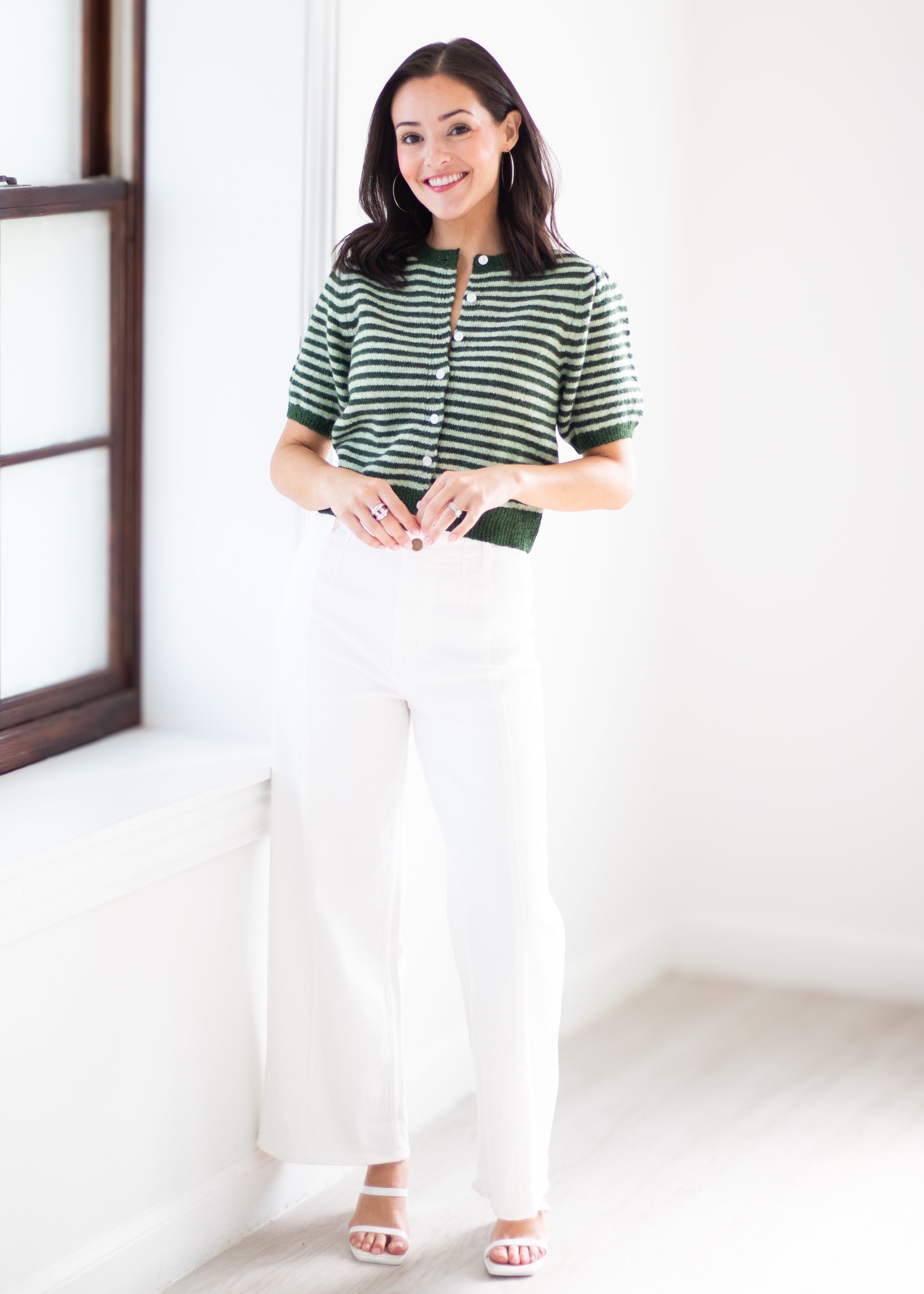 A woman modeling a green and white striped short-sleeve knit top with a split neckline, paired with white wide-leg pants and white sandals, standing in a bright white room.