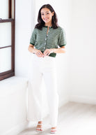 A woman modeling a green and white striped short-sleeve knit top with a split neckline, paired with white wide-leg pants and white sandals, standing in a bright white room.