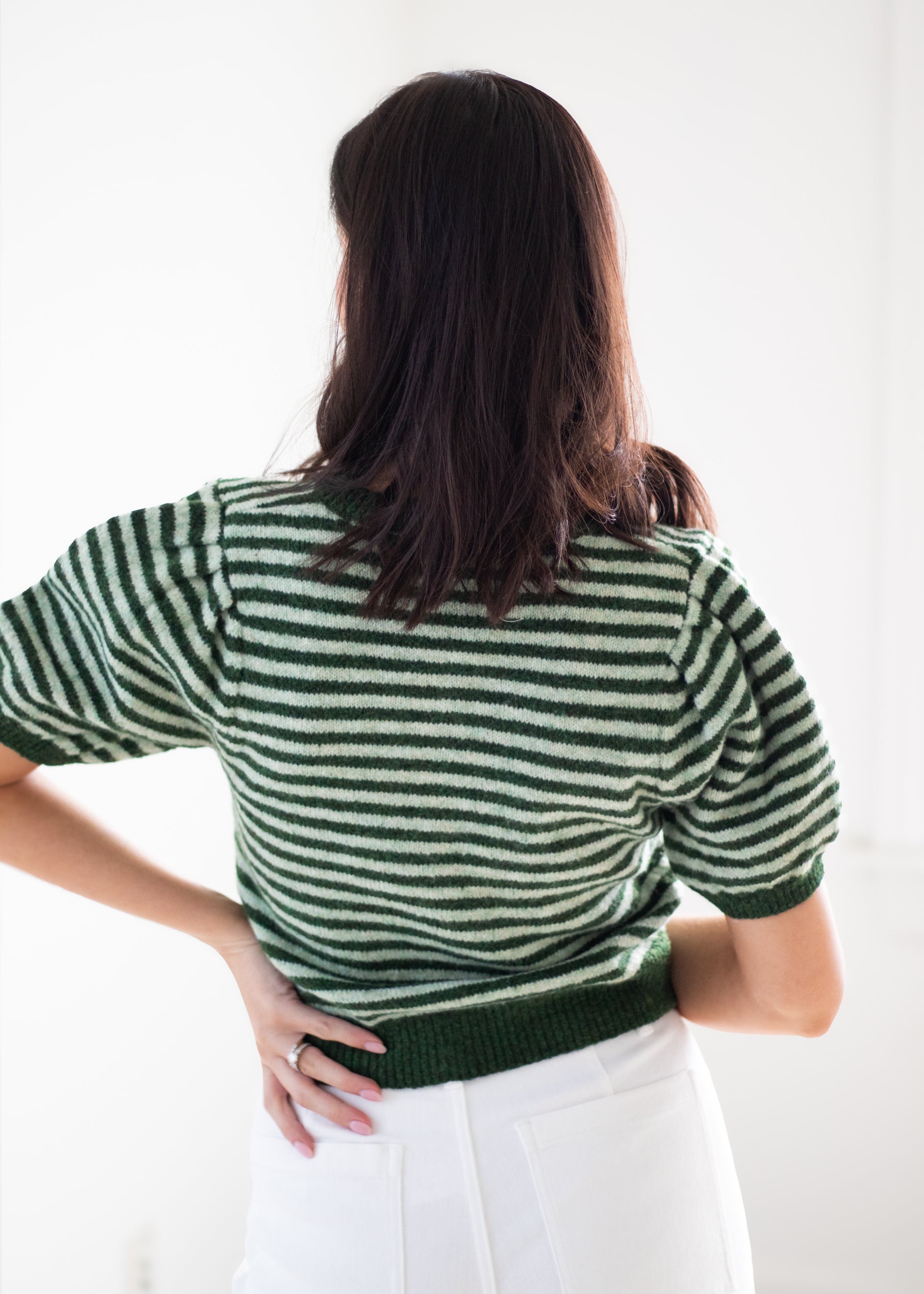 A woman shown from behind wearing a green and white striped short-sleeve knit top and white pants against a bright white background.