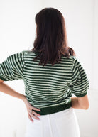 A woman shown from behind wearing a green and white striped short-sleeve knit top and white pants against a bright white background.