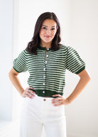 A woman modeling a green and white striped short-sleeve knit cardigan with white buttons, paired with white pants, against a bright white background.