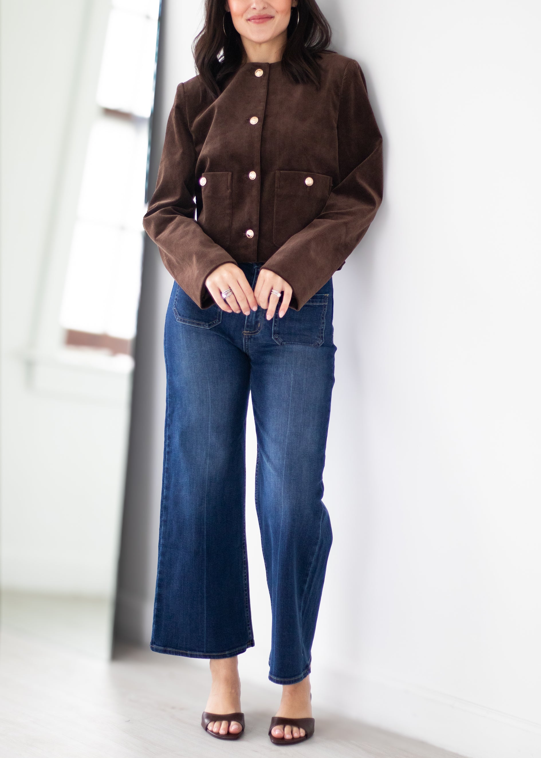 A woman wearing a brown corduroy jacket with gold-tone buttons and blue wide-leg jeans, standing in a bright indoor setting.