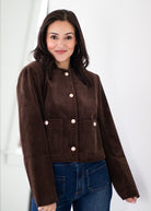 A woman wearing a cropped brown corduroy jacket with light pink buttons, paired with blue jeans, standing against a white background.