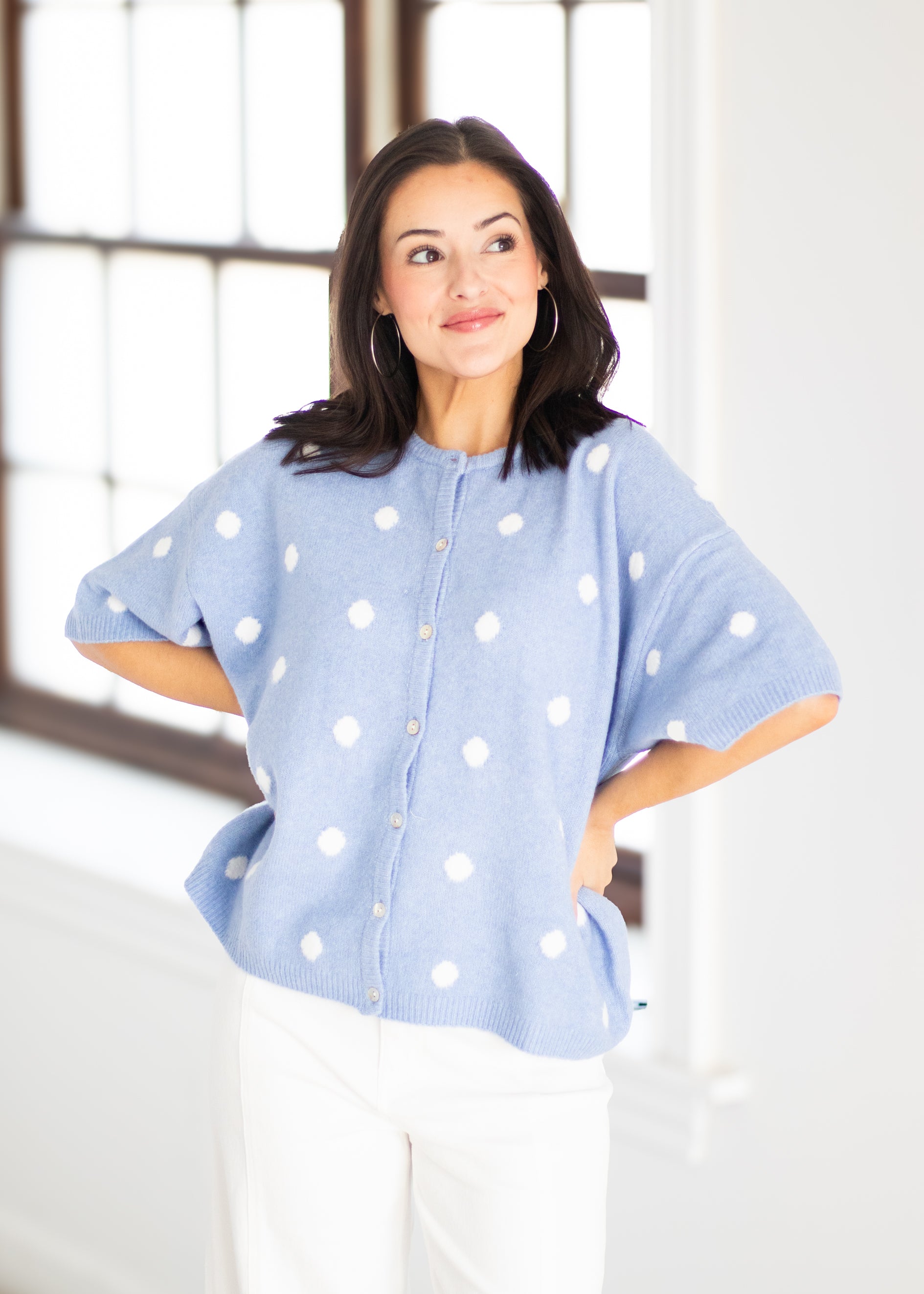 A woman modeling a light blue knit cardigan with white polka dots, paired with white pants, standing in a bright room with a large window.