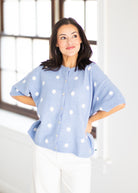 A woman modeling a light blue knit cardigan with white polka dots, paired with white pants, standing in a bright room with a large window.