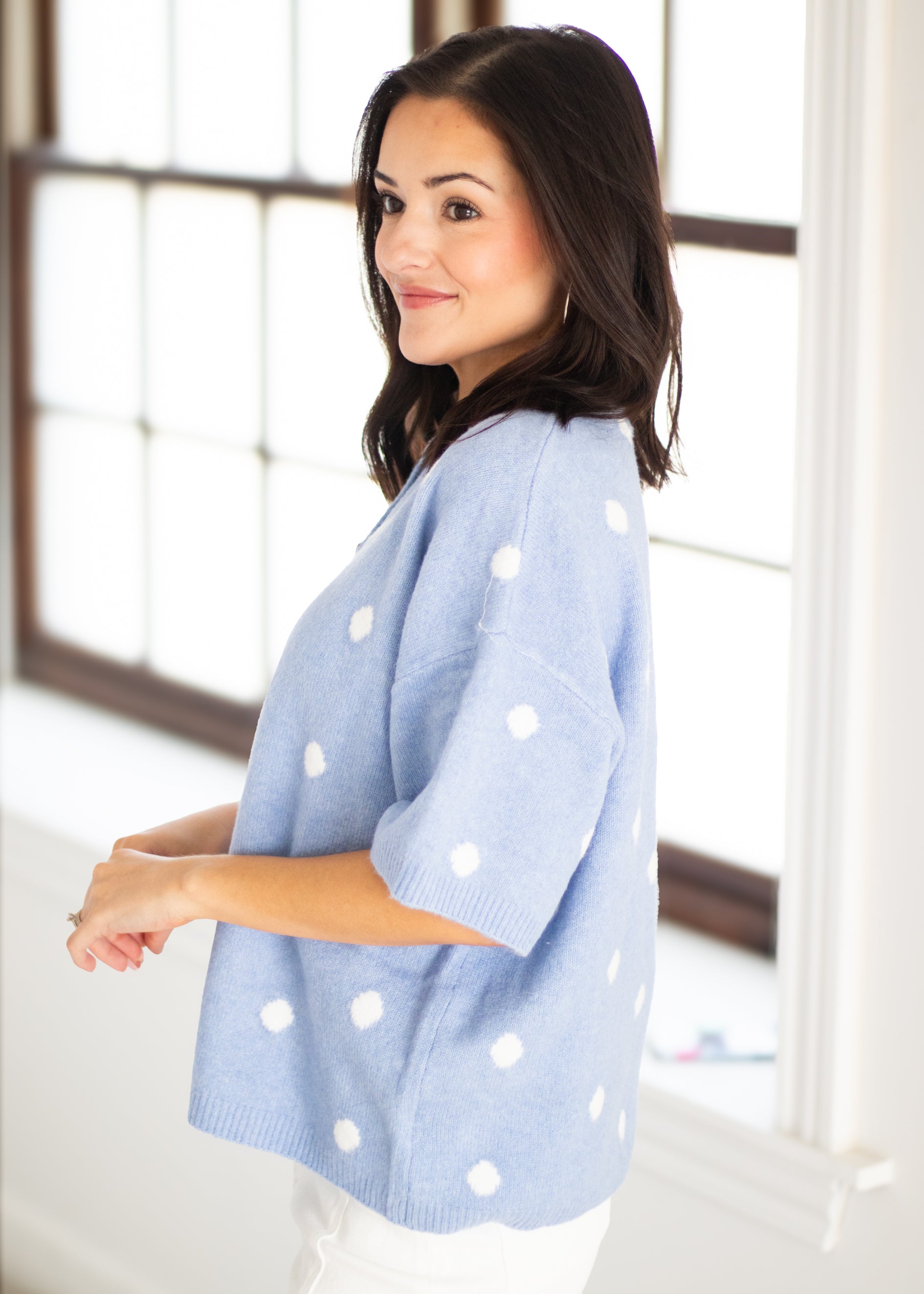 A woman wearing a light blue knit sweater with white polka dots, standing in a bright room with large windows.