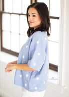 A woman wearing a light blue knit sweater with white polka dots, standing in a bright room with large windows.