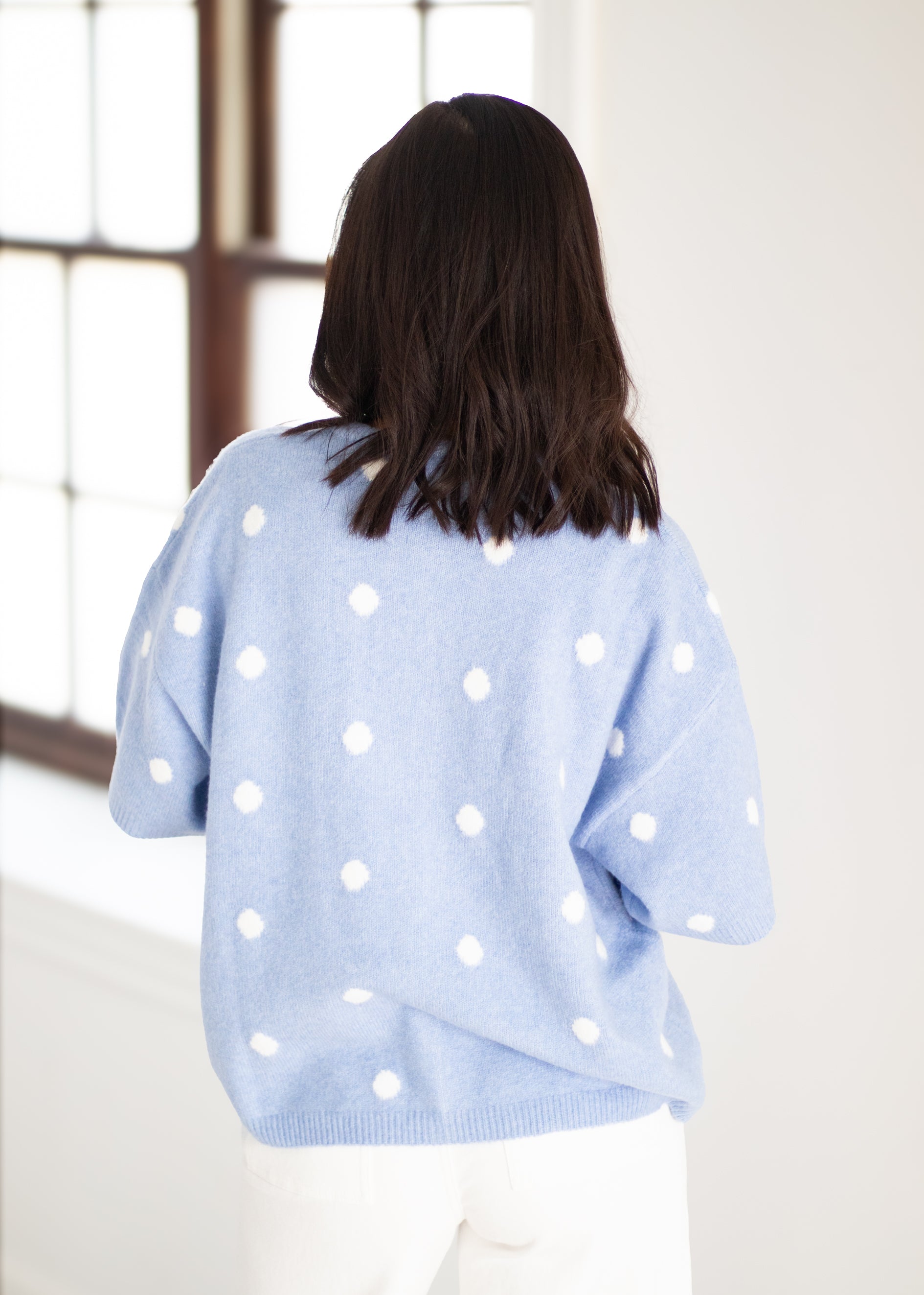 A woman viewed from behind wearing a light blue knit sweater with white polka dots, paired with white pants, standing in a bright indoor setting.