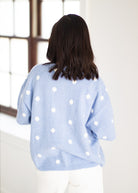 A woman viewed from behind wearing a light blue knit sweater with white polka dots, paired with white pants, standing in a bright indoor setting.