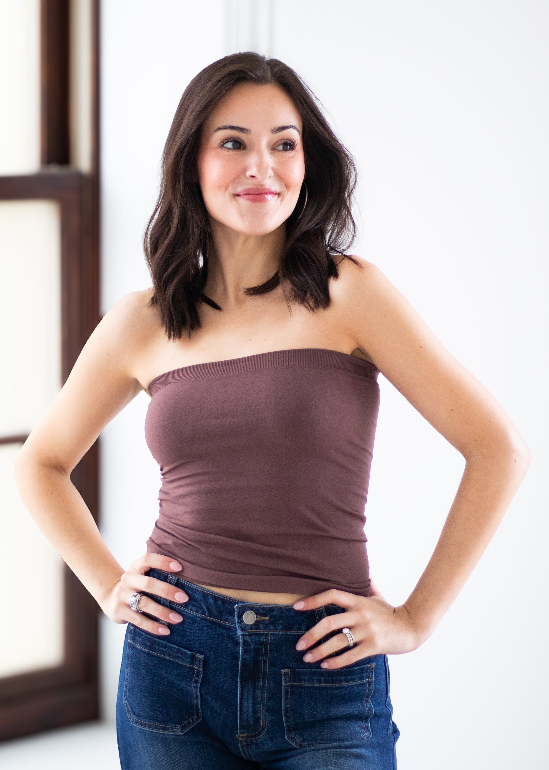 A woman with dark shoulder-length hair wearing a strapless mauve-brown tube top and blue jeans, posing with her hands on her hips against a bright white indoor background.