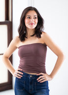 A woman with dark shoulder-length hair wearing a strapless mauve-brown tube top and blue jeans, posing with her hands on her hips against a bright white indoor background.