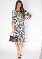 A woman modeling a black and white zebra-print shirt dress with three-quarter sleeves, holding a dark brown leather handbag against a plain white studio background.