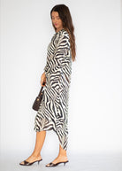 A woman modeling a long-sleeve midi dress with a bold black and white zebra print, styled with a small dark brown handbag and black mule sandals against a plain white studio background.