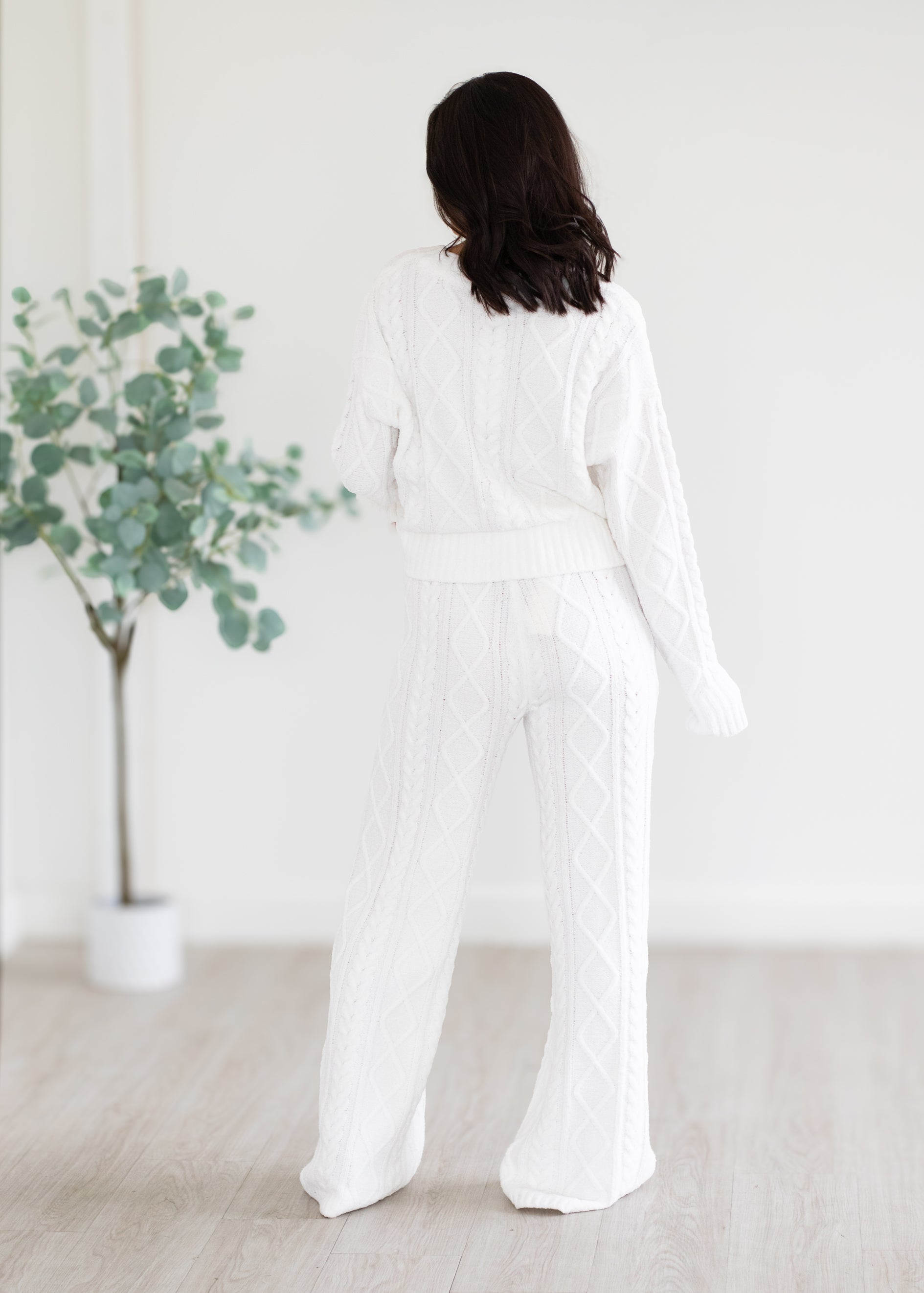 Person wearing a white outfit standing in a minimalistic room with a plant.