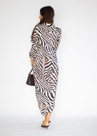 A woman is shown from behind wearing a long-sleeved zebra-print midi dress with a shawl collar. She is holding a small dark brown handbag and wearing black pointed-toe flats, set against a plain white studio background.