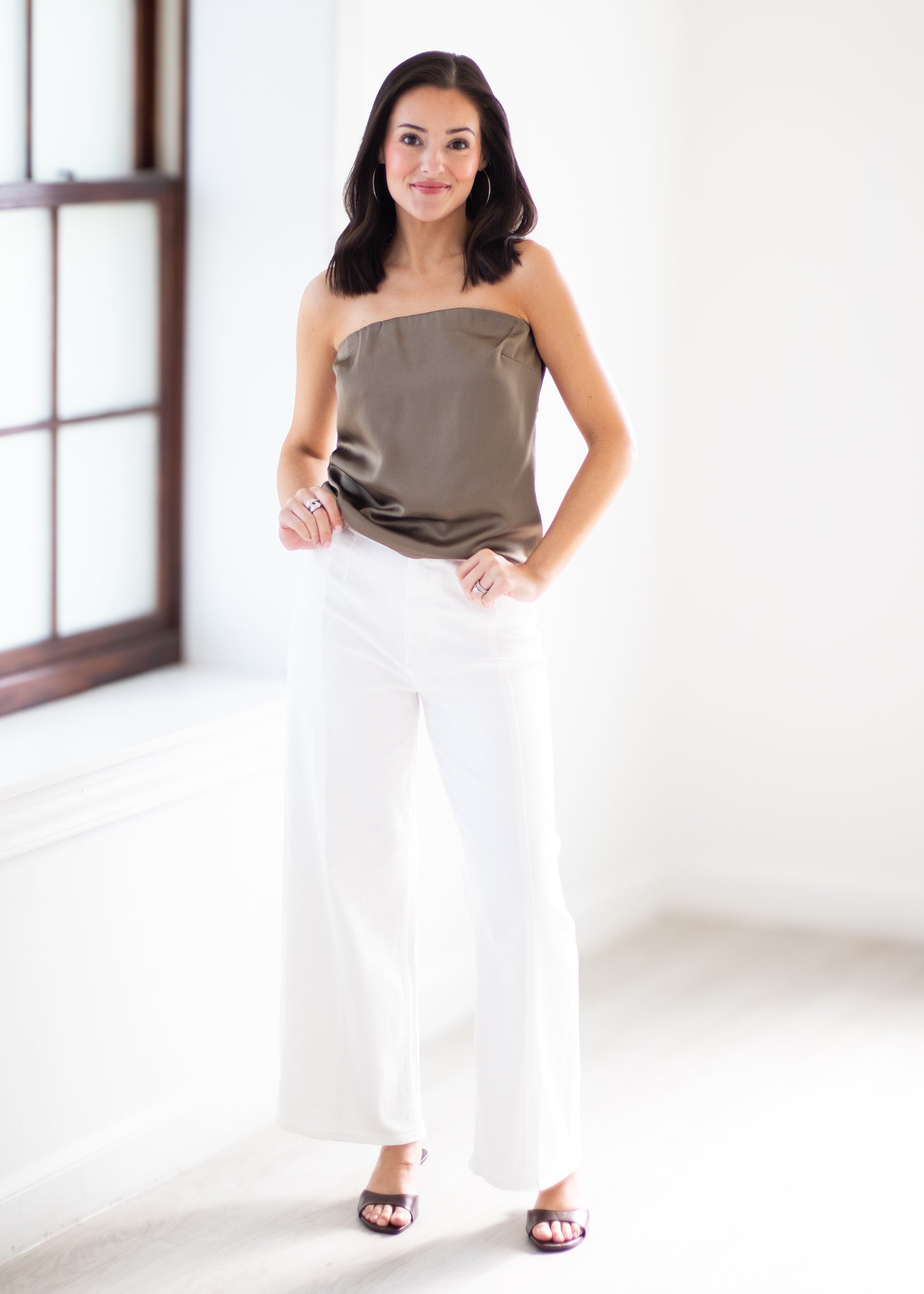 A woman modeling a strapless taupe satin top and white wide-leg pants in a bright, minimalist room with a window.