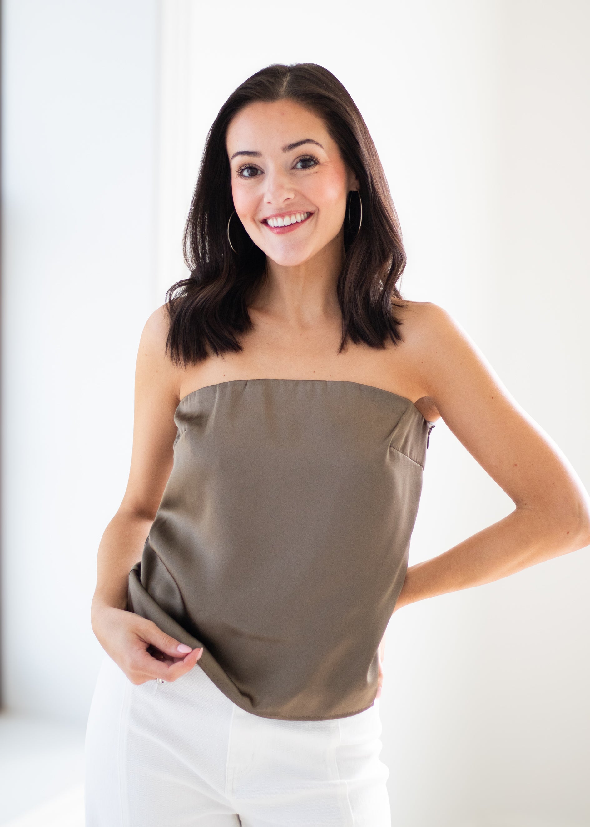 A woman wearing a strapless taupe top and white pants, smiling in a brightly lit indoor setting.