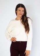 A woman modeling a cream-colored long-sleeve knit sweater paired with dark burgundy pants, standing against a plain light gray studio background.