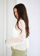 A woman with long, wavy brown hair is shown from a side-back angle, wearing a semi-sheer white long-sleeve blouse and olive green pants against a bright white background.