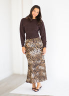 A woman modeling a dark brown long-sleeve knit top and a leopard-print maxi skirt, standing against a bright white studio background.