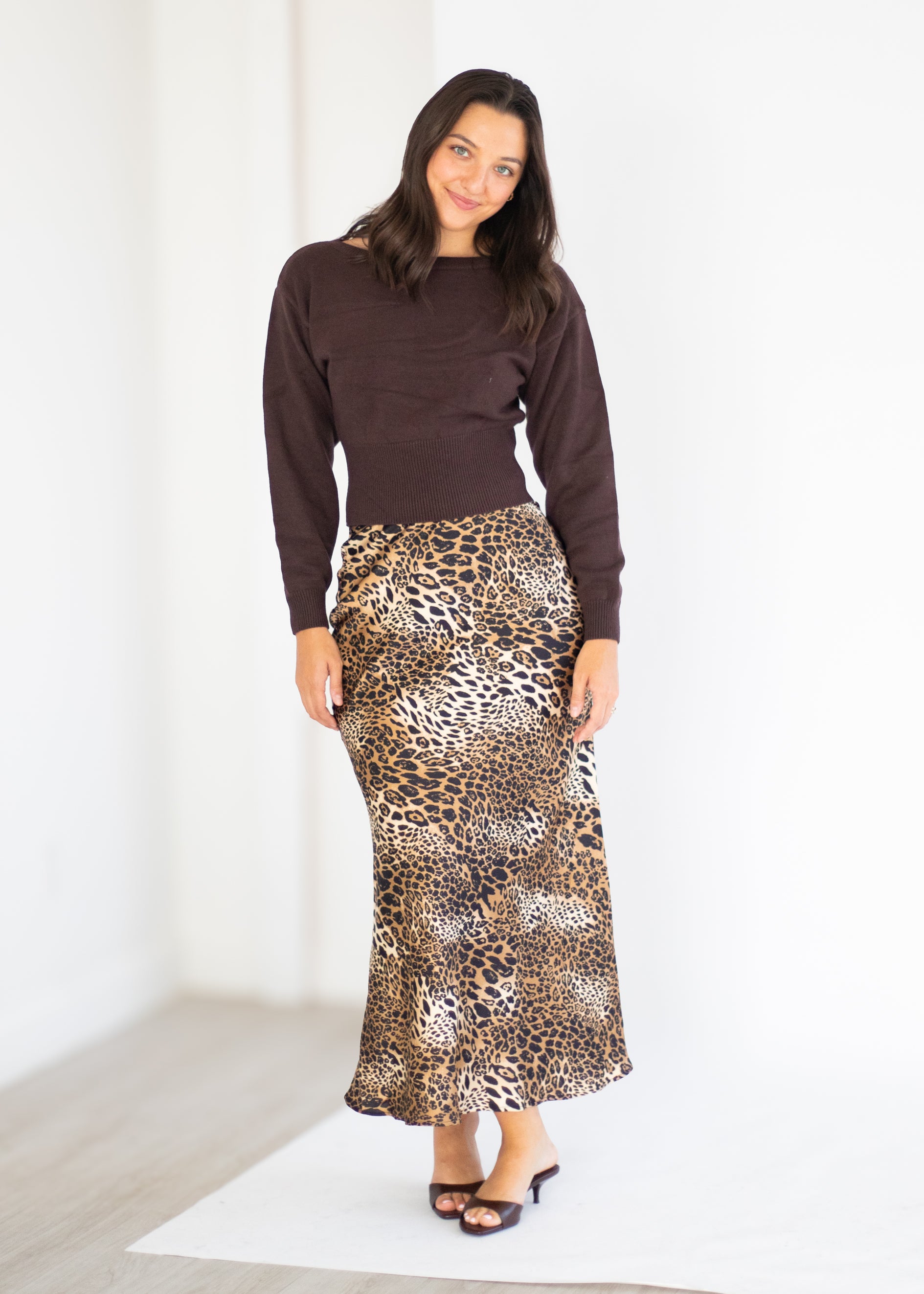 A woman modeling a dark brown long-sleeve knit top and a leopard-print maxi skirt, standing against a bright white studio background.