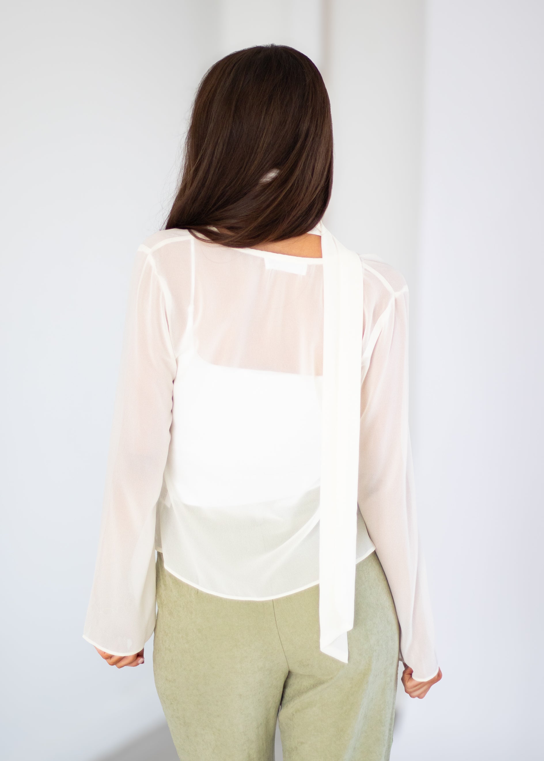 A woman viewed from behind wearing a white long-sleeve sheer blouse with a wide tie detail at the back, paired with light olive green pants, against a plain white background.