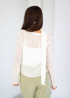 A woman viewed from behind wearing a white long-sleeve sheer blouse with a wide tie detail at the back, paired with light olive green pants, against a plain white background.