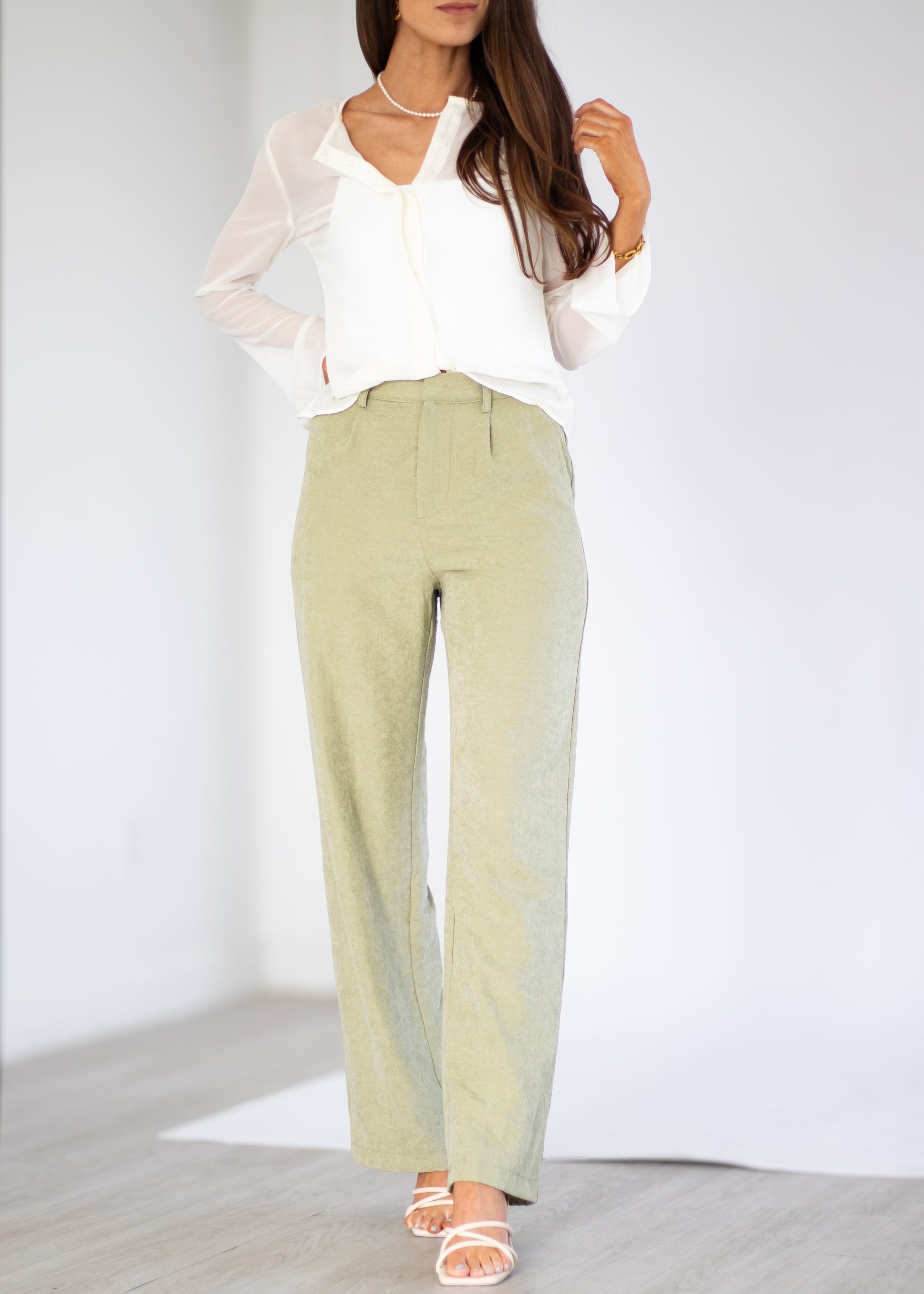 A woman modeling a white long-sleeve blouse and light sage green wide-leg trousers, paired with white strappy sandals, in a bright studio setting.