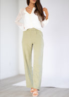 A woman modeling a white long-sleeve blouse and light sage green wide-leg trousers, paired with white strappy sandals, in a bright studio setting.