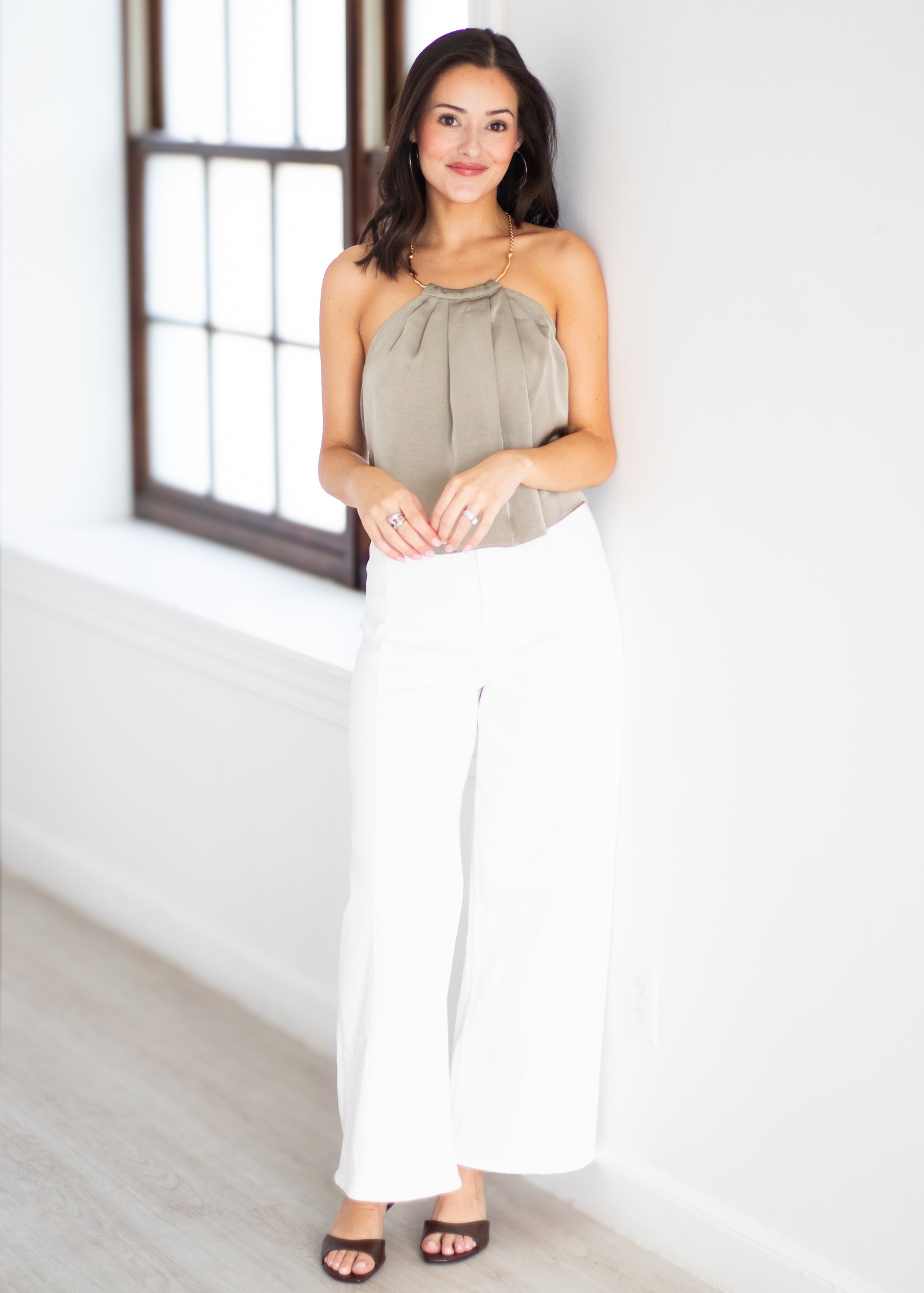 A woman modeling a sleeveless taupe halter top and white wide-leg pants, standing in a bright white room with a window.