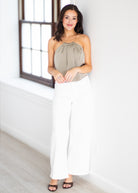 A woman modeling a sleeveless taupe halter top and white wide-leg pants, standing in a bright white room with a window.