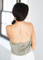 A woman shown from behind wearing a gold chain necklace with a long drop tassel, paired with a light sage green strapless top and white bottoms, standing in a bright indoor setting.