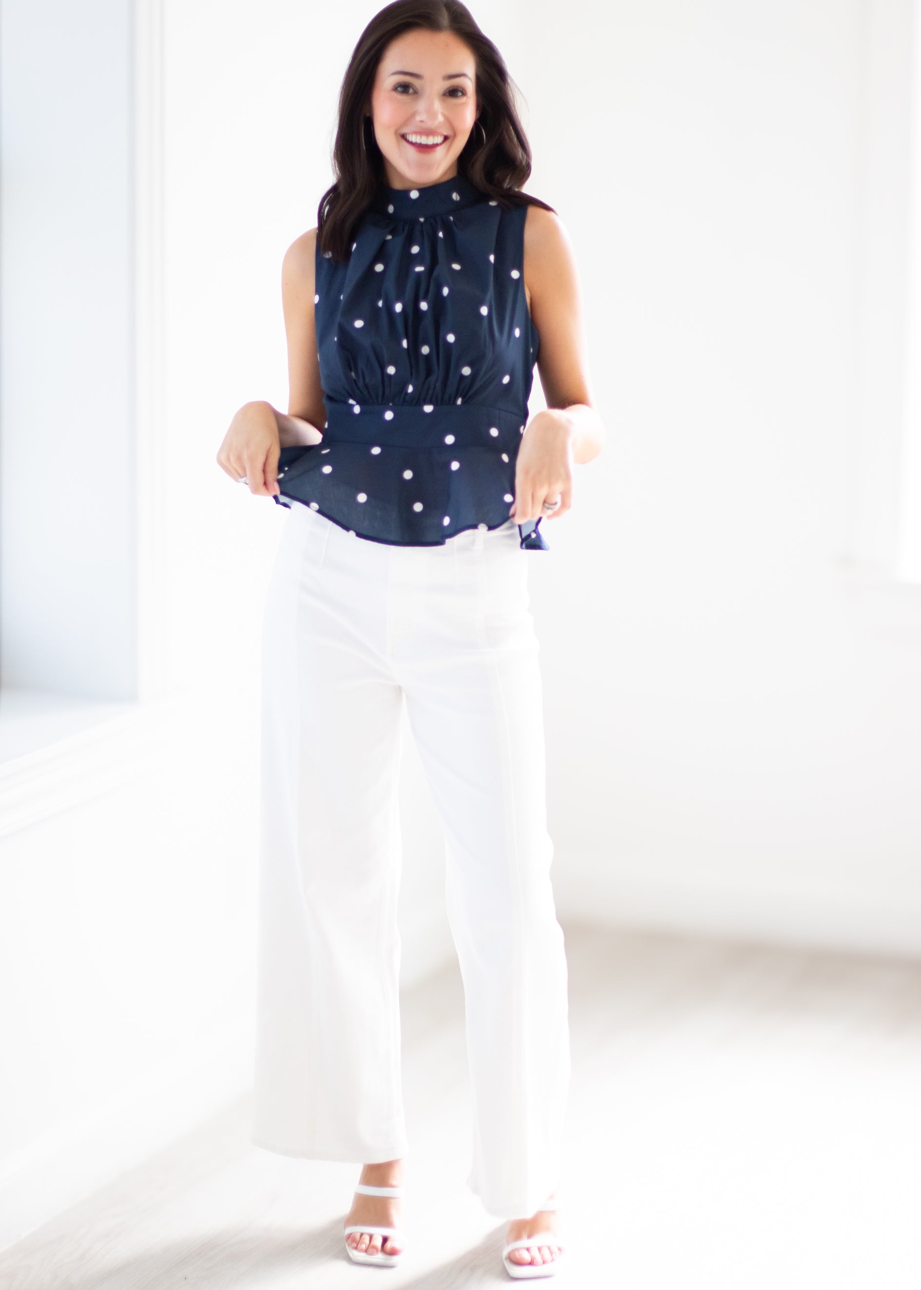 A woman modeling a sleeveless navy blue blouse with white polka dots and white wide-leg pants in a bright, minimalist indoor setting.
