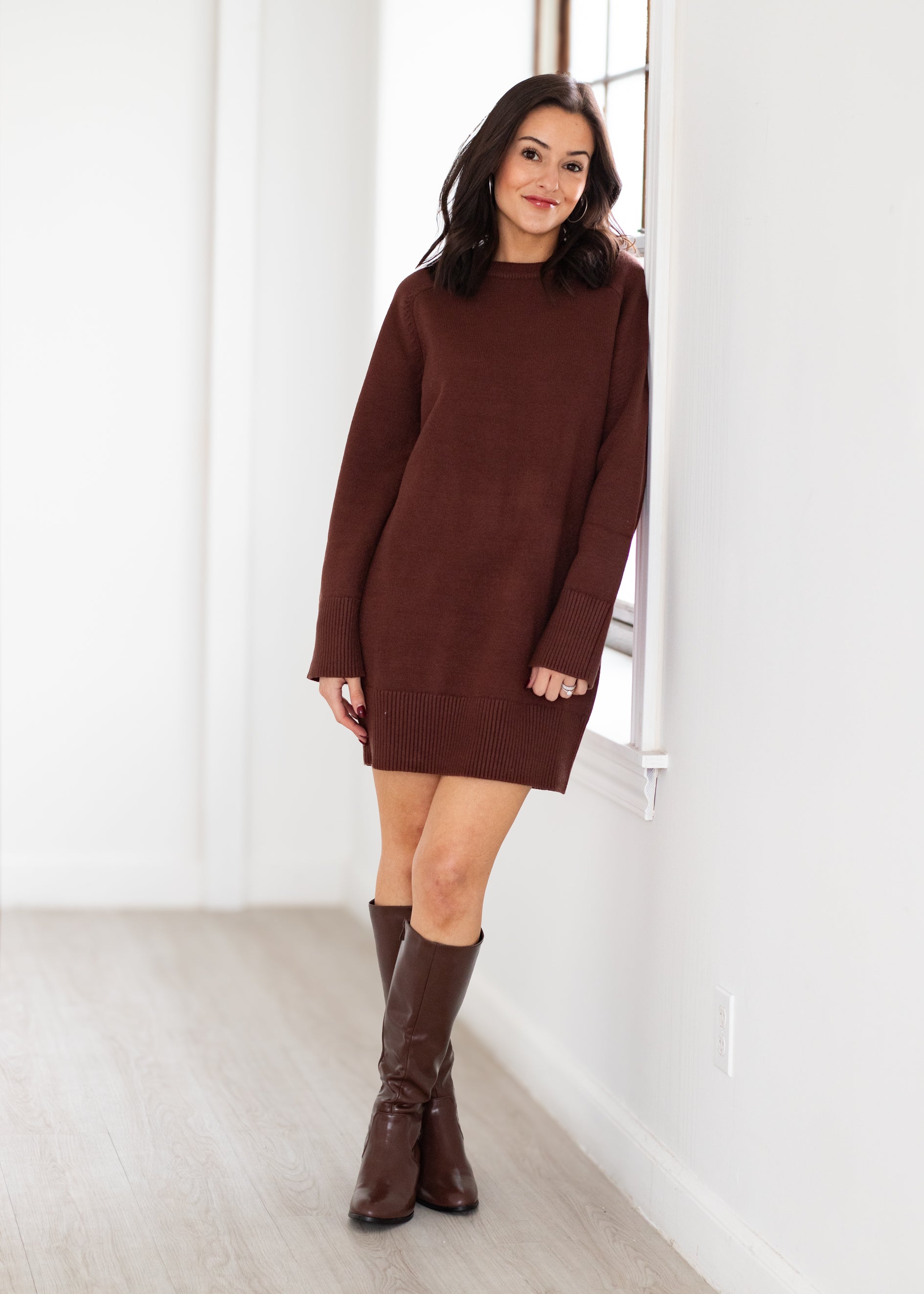 Woman wearing a brown sweater dress and knee-high boots in a minimalistic room.
