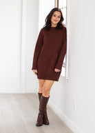 Woman wearing a brown sweater dress and knee-high boots in a minimalistic room.