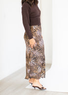 A woman wearing a brown long-sleeve knit top and a leopard-print maxi skirt, paired with dark heeled sandals, standing in a bright indoor setting.