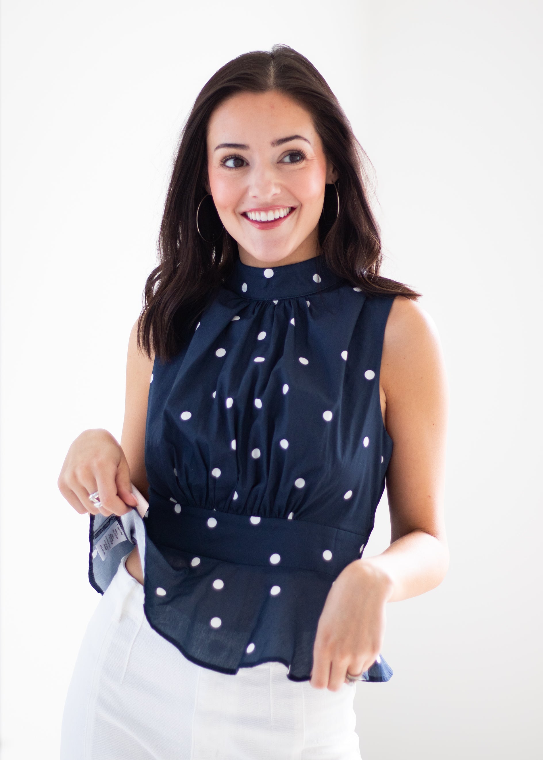 A smiling woman wearing a sleeveless navy blue blouse with white polka dots and a gathered waist, paired with white pants, against a plain white studio background.