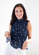 A smiling woman wearing a sleeveless navy blue blouse with white polka dots and a gathered waist, paired with white pants, against a plain white studio background.