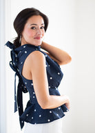A woman wearing a navy blue sleeveless top with white polka dots and a tie-neck detail, paired with white pants, photographed against a bright white background.