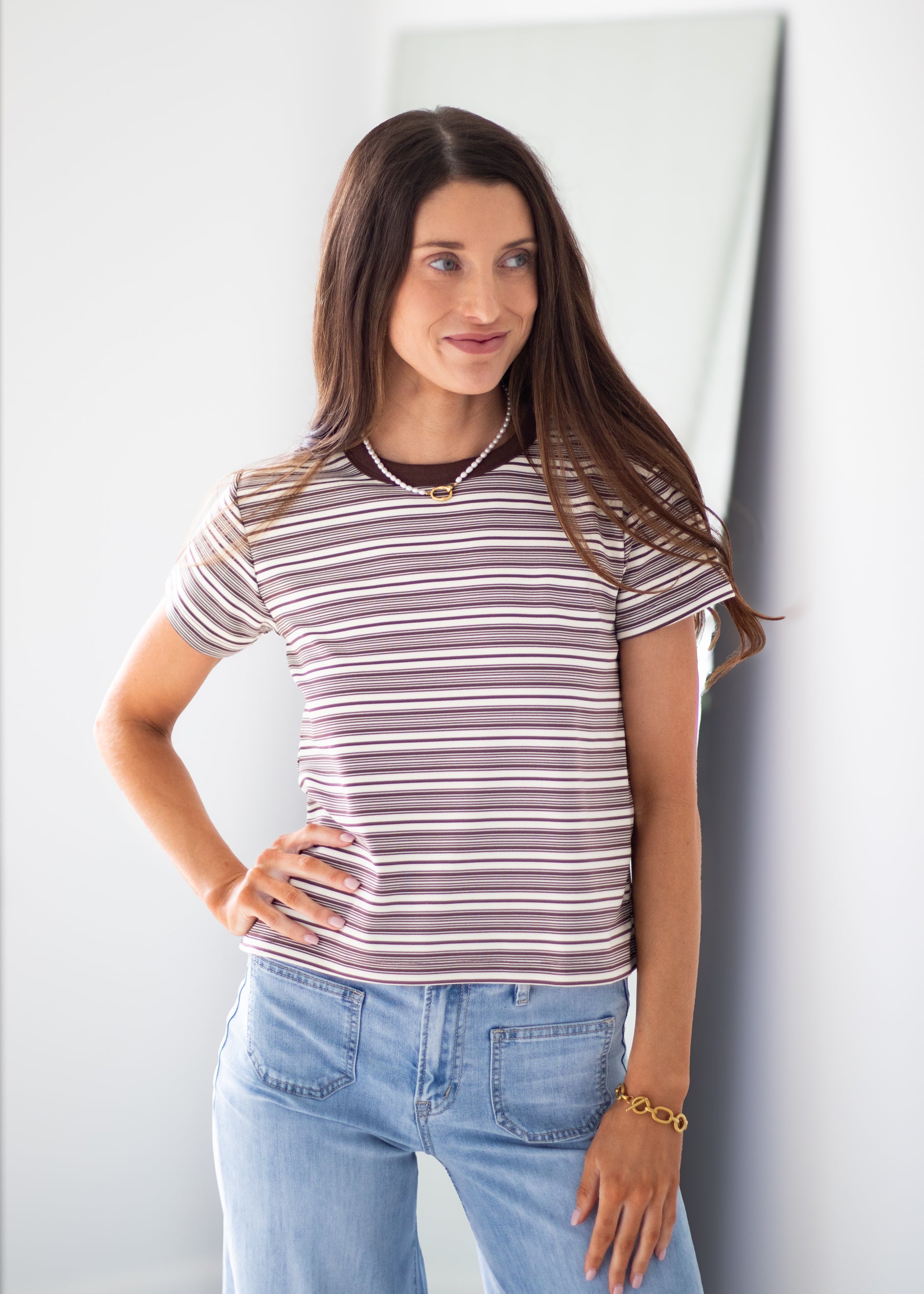 A woman wearing a short-sleeve crewneck t-shirt with thin brown and white horizontal stripes, paired with light blue jeans, standing against a light gray wall.