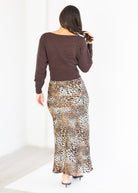 A woman shown from behind wearing a dark brown long-sleeve knit top and a leopard-print maxi skirt, standing in a bright white studio setting.