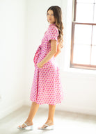 Woman wearing a pink patterned dress standing in a bright room with a window.