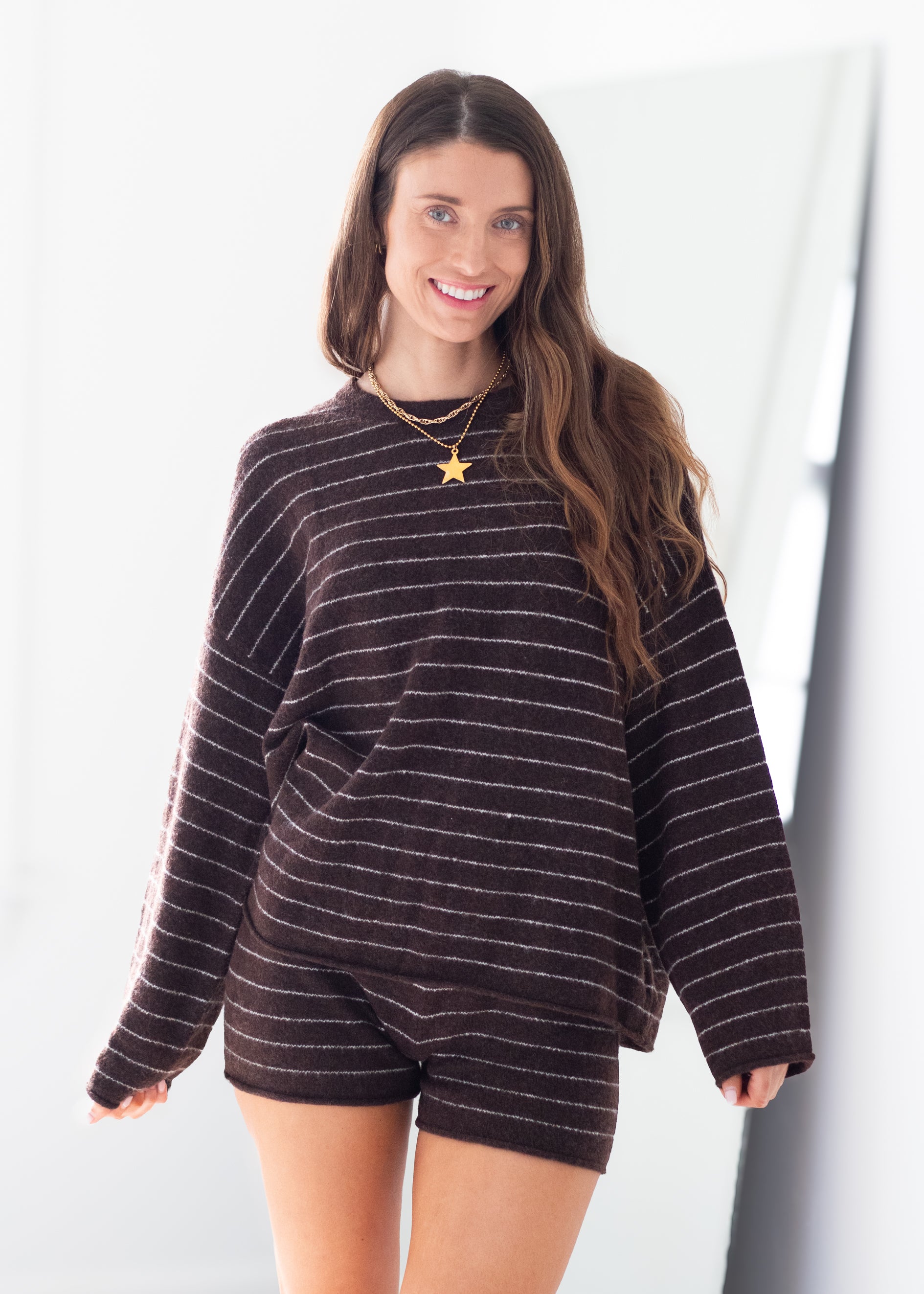 A woman modeling a dark brown and white striped knit lounge set consisting of a long-sleeve top and matching shorts, accessorized with gold necklaces.