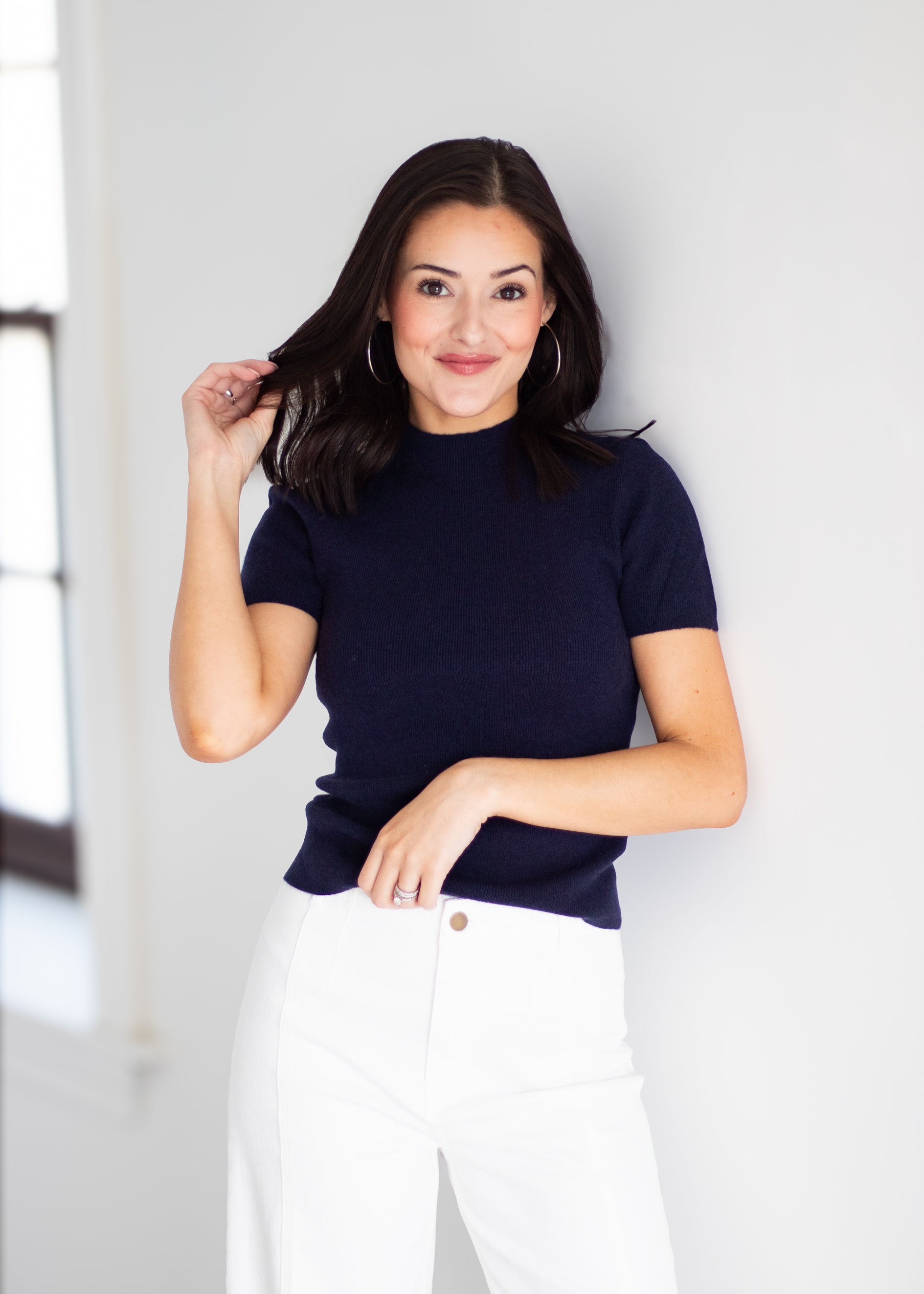 A woman modeling a short-sleeve mock neck knit top in navy blue, paired with white pants, standing against a light gray wall.