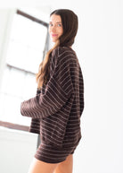 A woman wearing an oversized brown knit sweater with thin white stripes, photographed in a bright indoor setting.