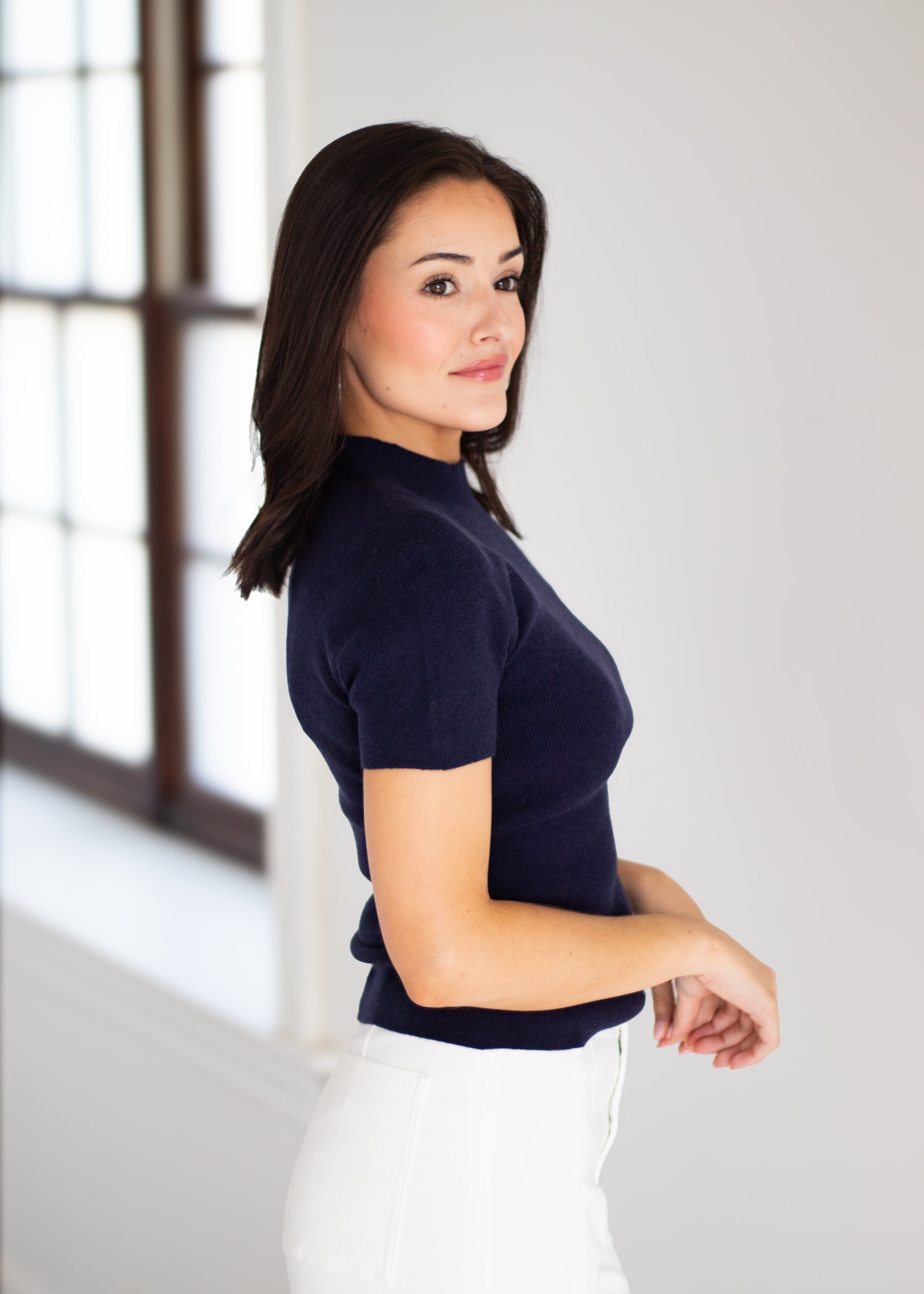 A woman with dark hair is shown from the side, wearing a short-sleeve navy blue mock neck knit top and white pants, standing in a brightly lit room with a window in the background.