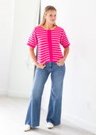 A woman modeling a short-sleeve knit top with bright pink and white horizontal stripes, featuring a solid pink collar and button placket, paired with blue flared jeans and white shoes.
